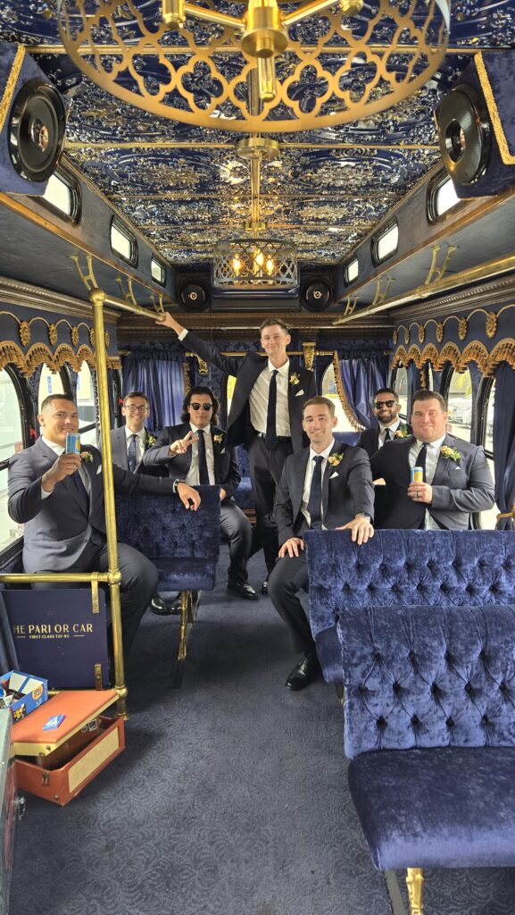 A group of groomsmen in navy suits celebrating inside a luxury Parlor Car Trolley with navy velvet seating and ornate gold ceilings.