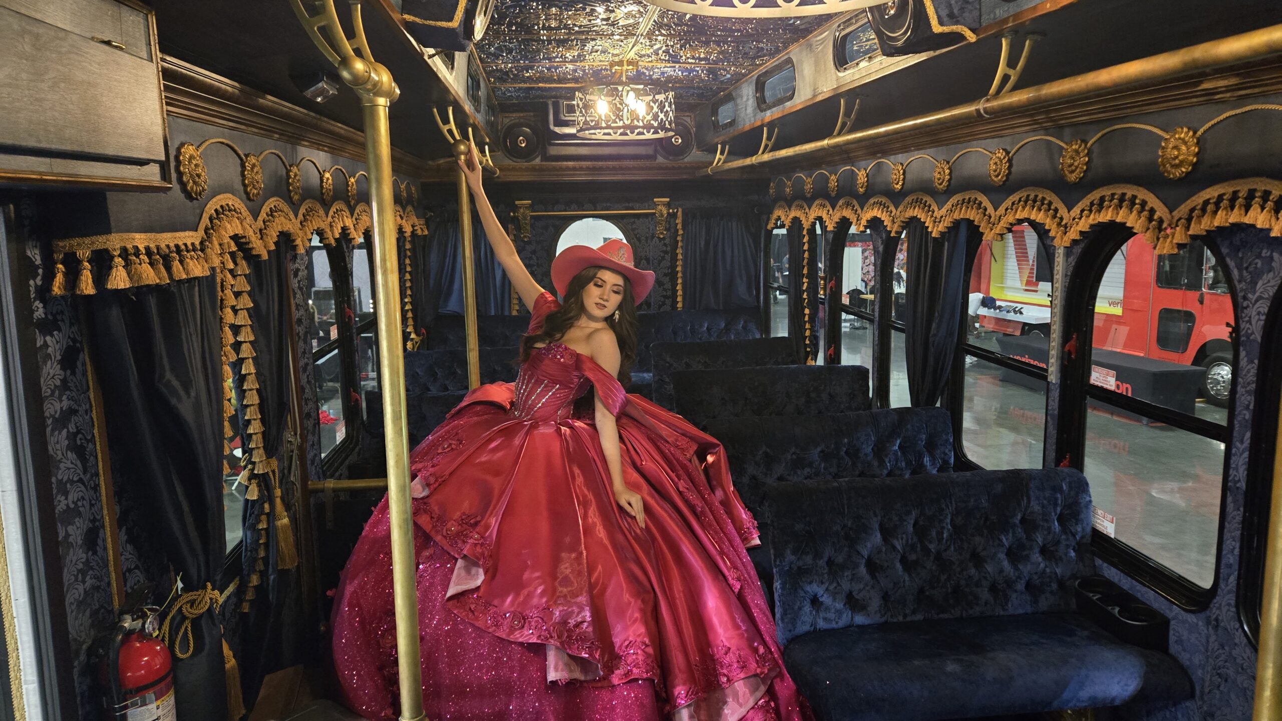 A woman in a voluminous pink gown and cowboy hat posing inside the Parlor Car Trolley featuring blue velvet seating and gold ornate trim.