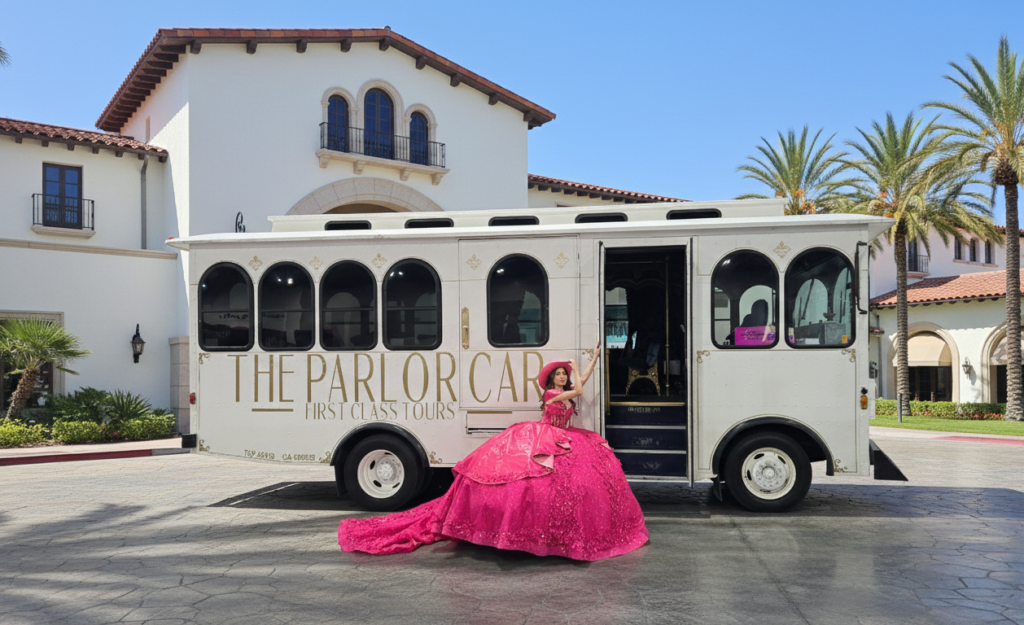 A young woman in a bright pink Quinceañera dress posing by the entrance of a white Parlor Car Trolley in front of a luxury Spanish-style hotel.